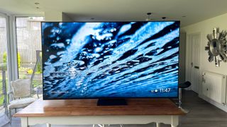 The TCL 85C805K 4K TV on a table in a room with a garden visible through a window in the background. On screen is a close-up of a water surface.